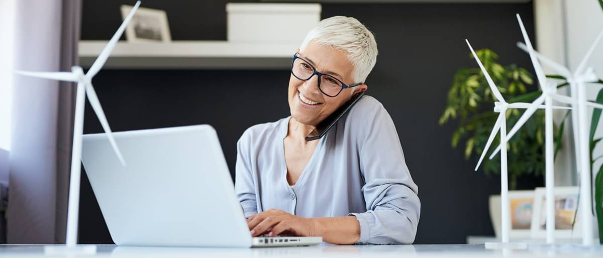 Attraktive ältere Frau am Telefon sprechend und auf Laptop schauen, während sie im Büro sitzt. Nachhaltigkeitskonzept
